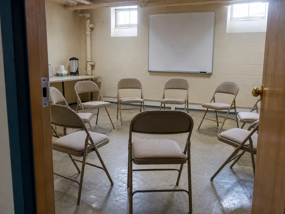 A small group counseling room with chairs set in a circle, seen through the open door, morning light, coffee urn on a side table