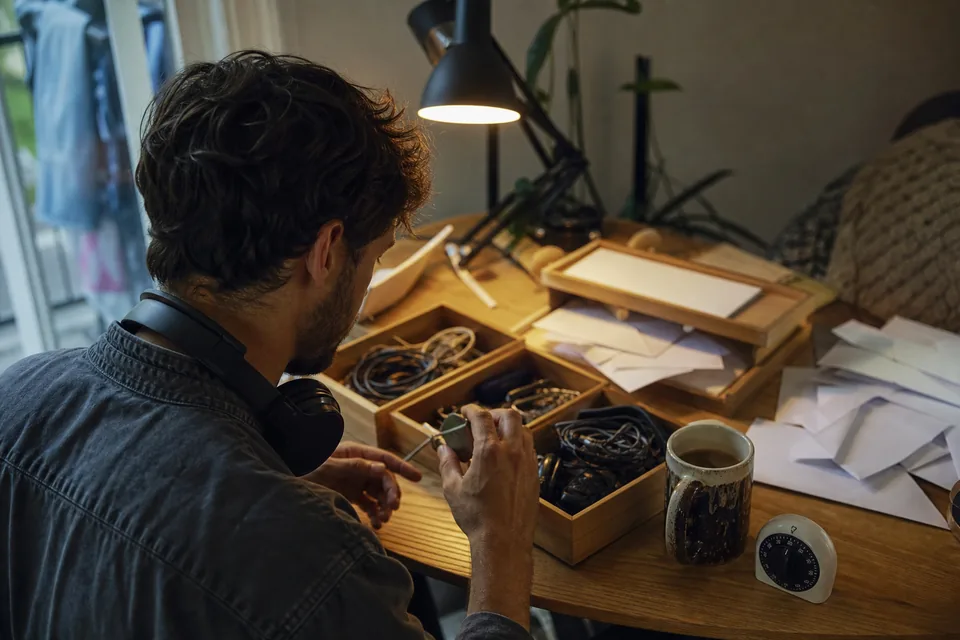 A man at a busy home desk, headphones around his neck, sorting a pile of things into trays