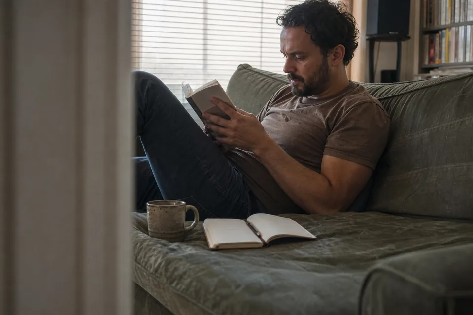 A person reading on a sofa with a cup of tea and a notebook, afternoon light