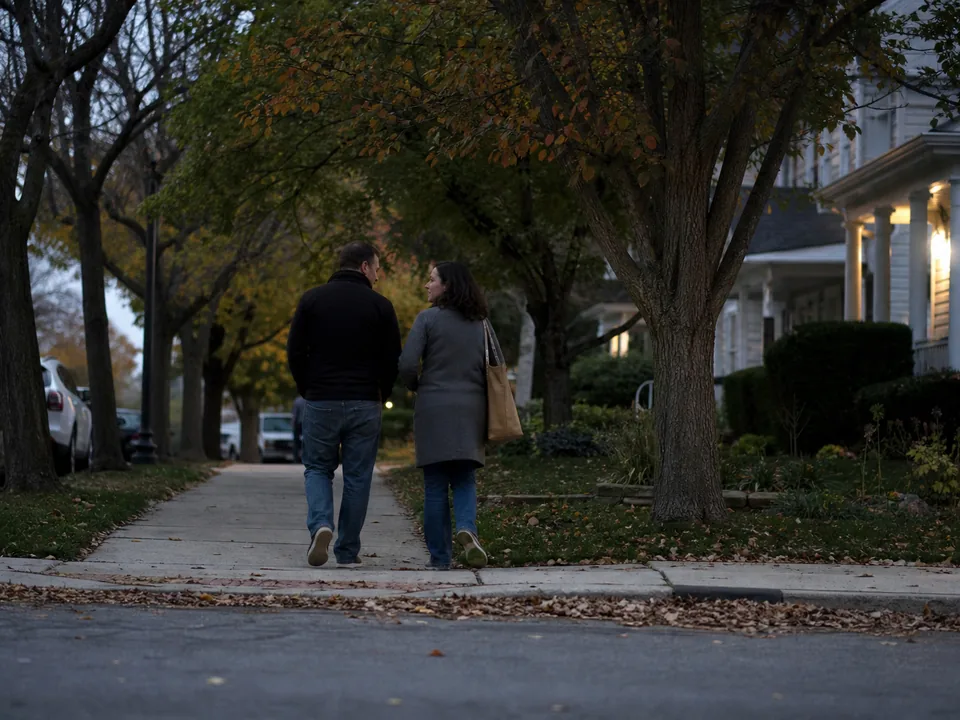 A couple walking side by side on a residential street at dusk, seen from behind, porch lights on