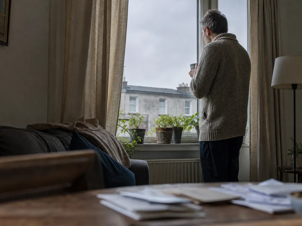 A man standing at a living room window with a mug, morning light on the curtains, a sofa with a folded blanket in the foreground