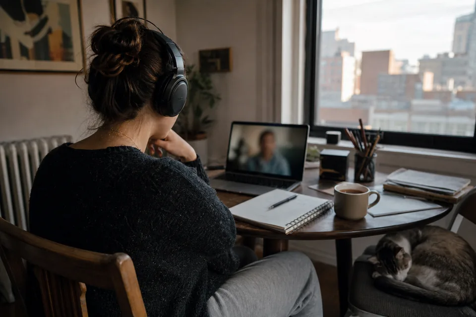 A woman at a small table at home on a video call on her laptop, headphones on, seen from behind over her shoulder, a cat on the chair beside her