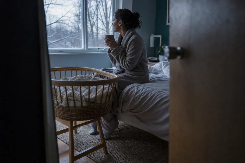A mother sitting on the edge of a bed at dawn with a mug, a bassinet beside the bed